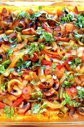 A traybake taken from above. Mixed vegetables including sliced red peppers and onions are garnished with fresh green herbs.