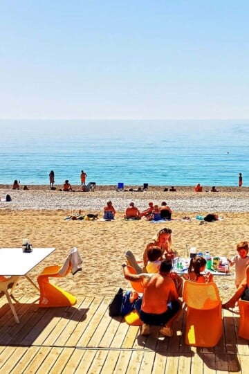 A family sits at a table on a raised wooden terrace on Ölüdeniz Beach. People are sat on the beach in the background and the sea is flat calm.