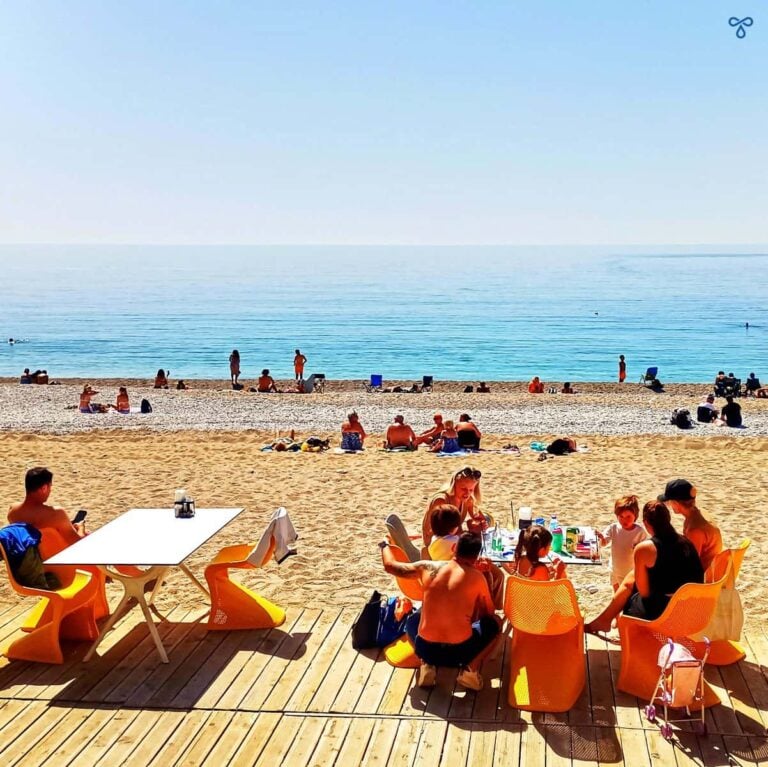 A family sits at a table on a raised wooden terrace on Ölüdeniz Beach. People are sat on the beach in the background and the sea is flat calm.