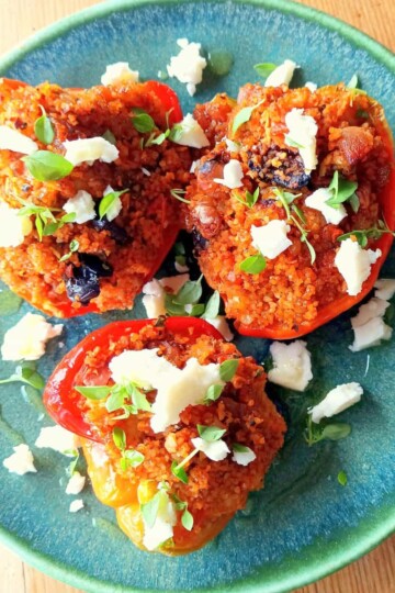A square image of three halved peppers stuffed with bulgur wheat. The photo is taken from above and the peppers are garnished with crumbled white cheese and torn basil leaves.