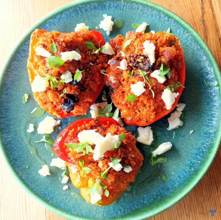A square image of three halved peppers stuffed with bulgur wheat. The photo is taken from above and the peppers are garnished with crumbled white cheese and torn basil leaves.