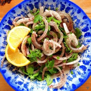 A sliced onion salad coasted in sumac and chopped parsley. Two slices of lemon are on the side of the bowl.