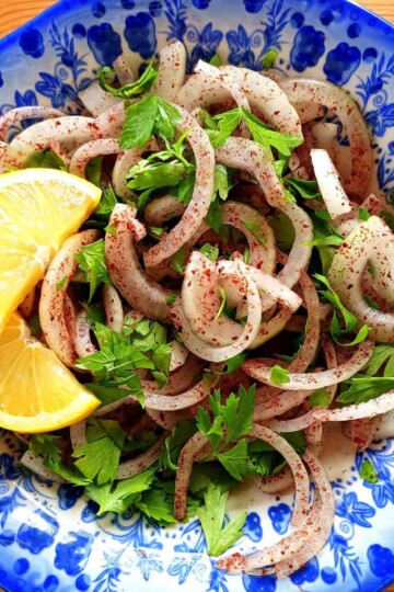 A sliced onion salad coasted in sumac and chopped parsley. Two slices of lemon are on the side of the bowl.