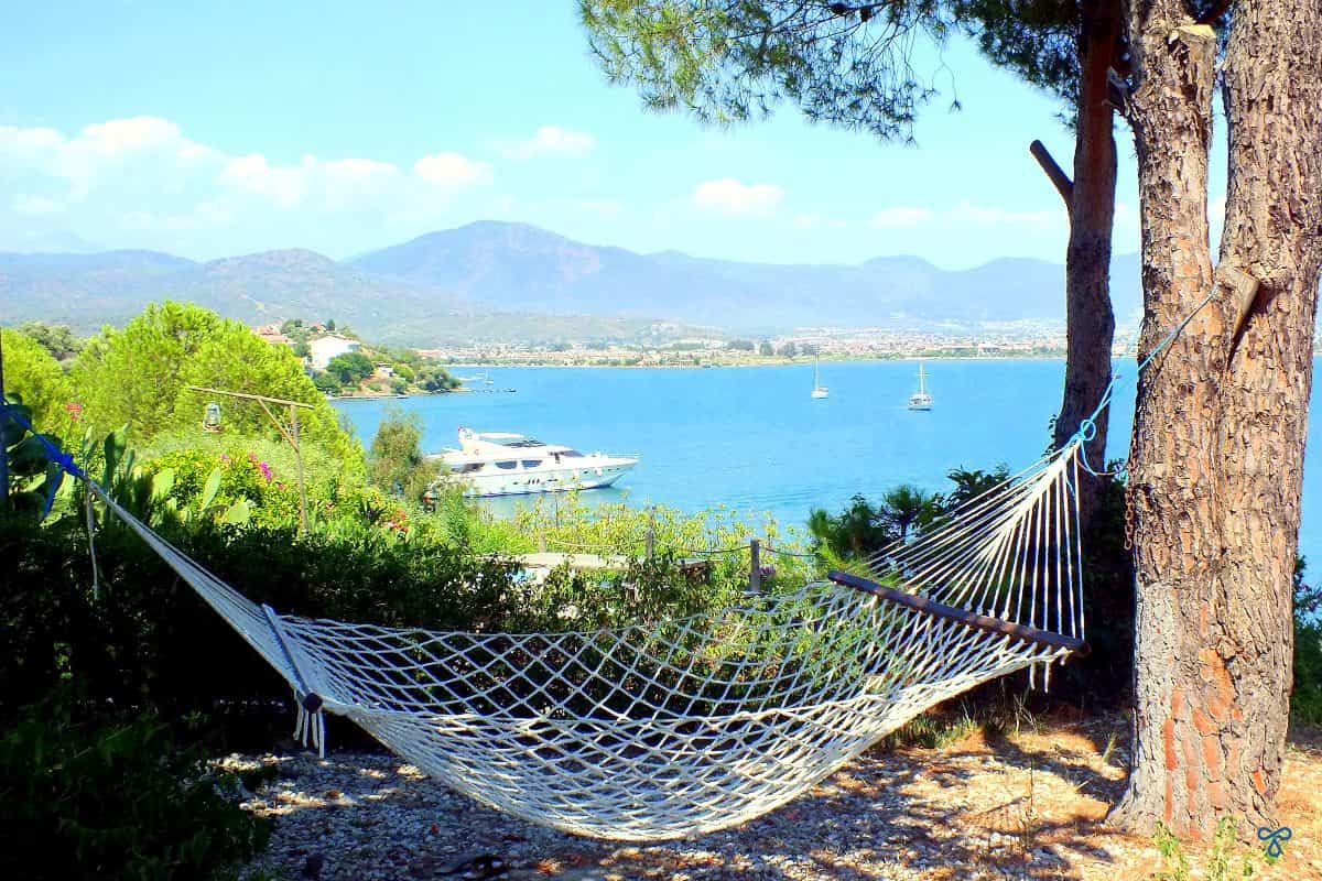 A woven hammock hanging between two tree trunks. The sea and mountains are in the background.