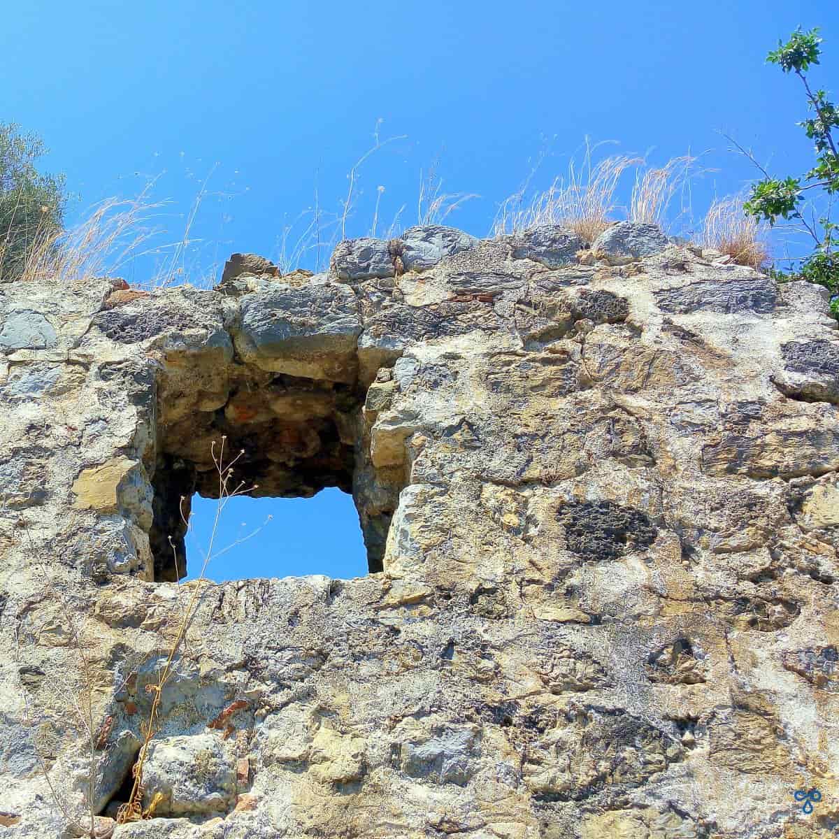 An ancient stone wall with small window opening revealing clear blue sky. Weeds are growing from the top of the wall.