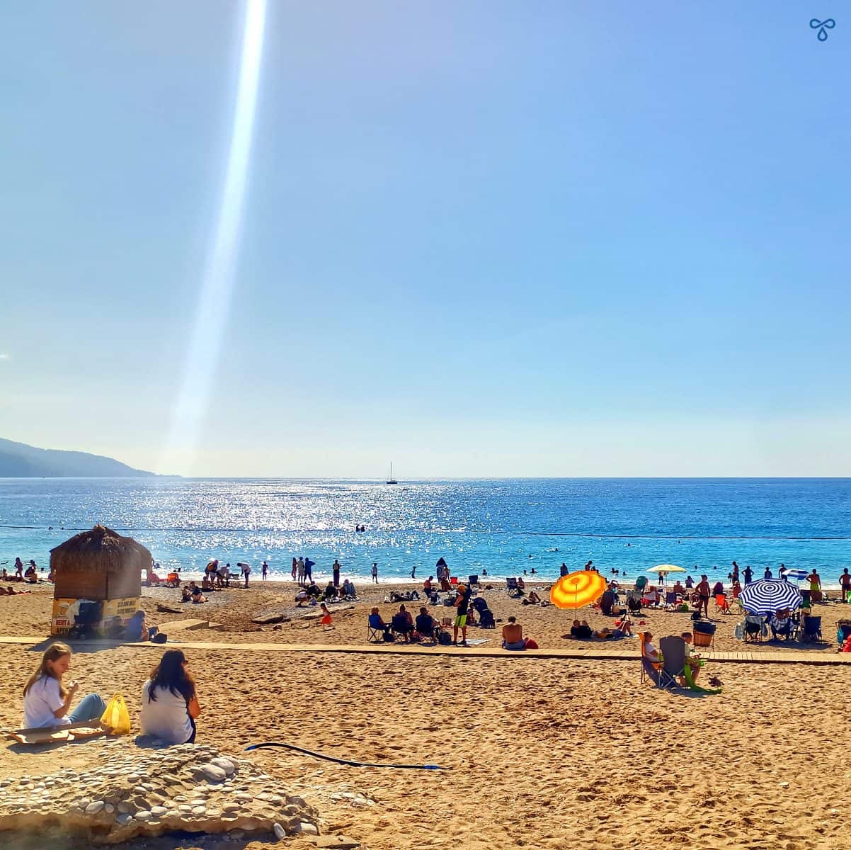 People sitting along Belcekiz Beach in Ölüdeniz. The sky is cloudless.