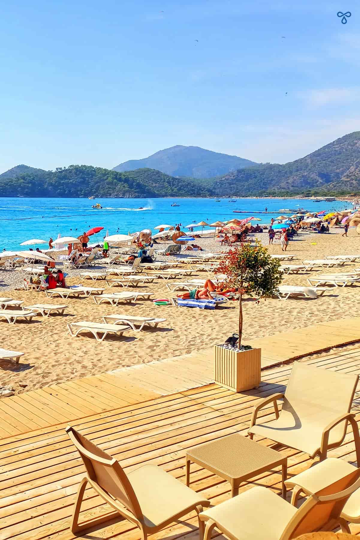 A wooden deck with tables and chairs on Belcekiz Beach in Ölüdeniz. Sun loungers and the sea are in the background.