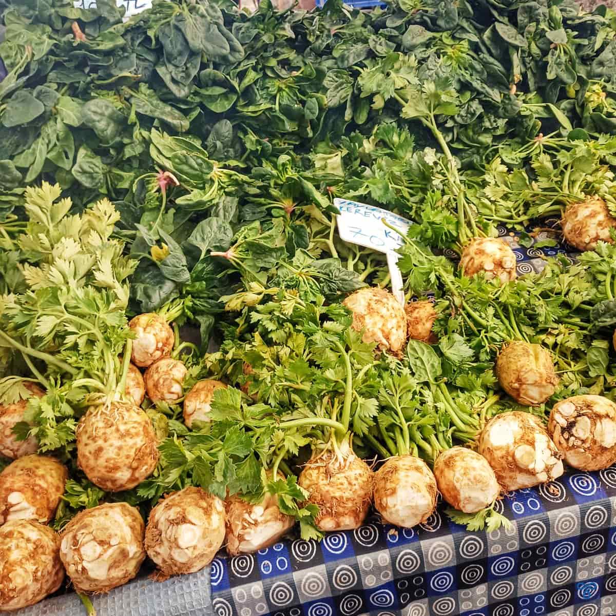 Bulbs of celeriac with their stems & leaves intact are piled high on a market stall. Spinach is stacked on top.