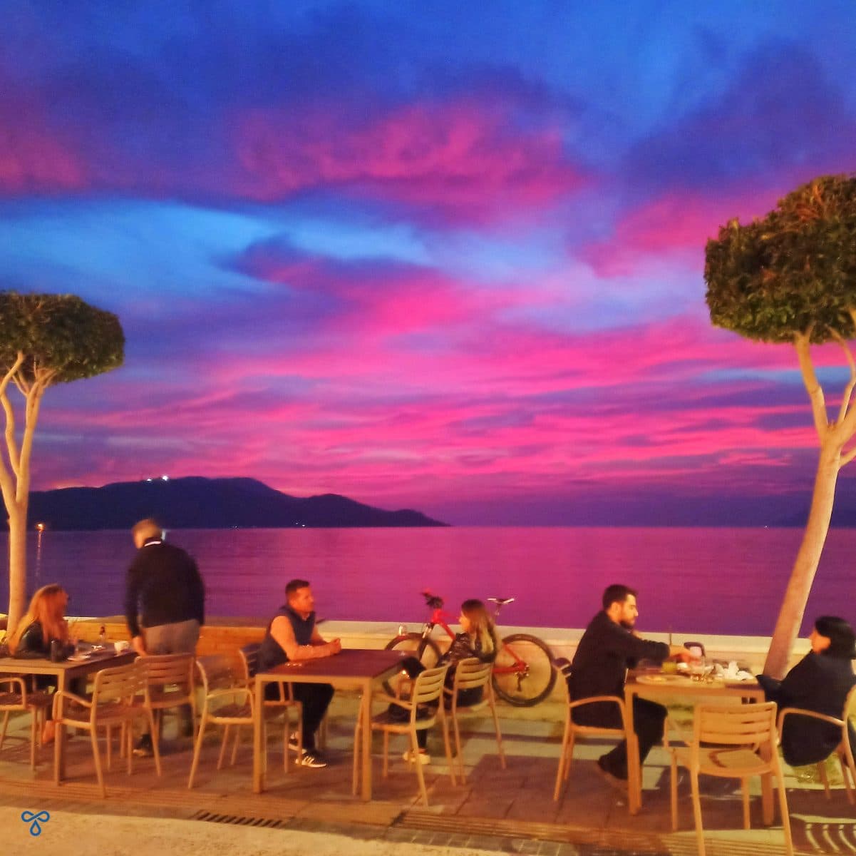 People sat at tables along Çalış Beach seafront. There is a tree either side and a fuschia sunset.