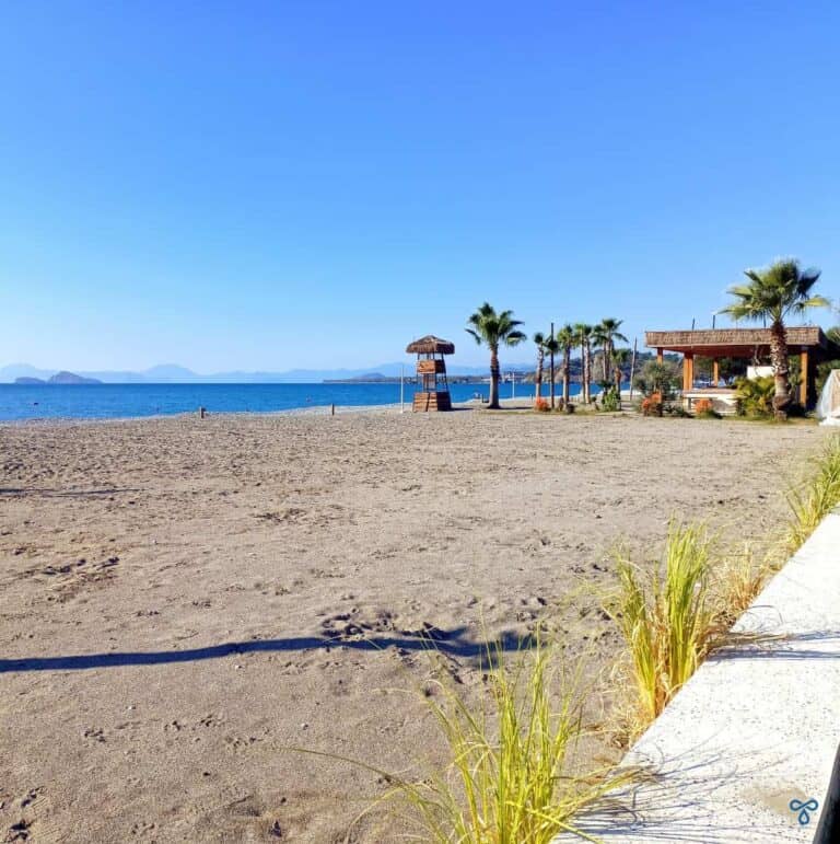 Expanse of beach with a wooden beach bar and palm trees in the distance at Jiva in Çalış.
