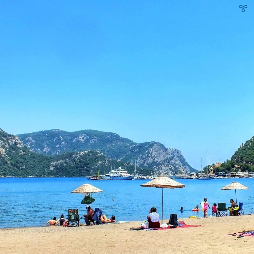 People sitting on the sand at İçmeler. They are sitting under rattan parasols. Hills back the bay.