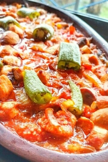 A close up of a shallow oval clay pot filled with Turkish chicken casserole. It is topped with chunks of green pepper.