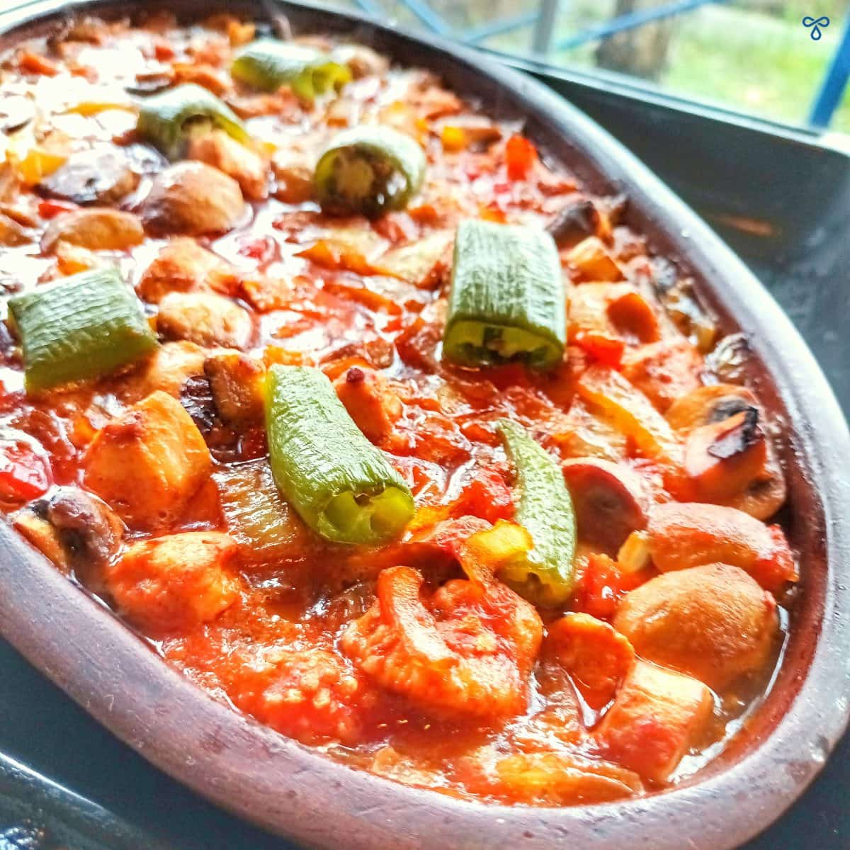 A close up of a shallow oval clay pot filled with Turkish chicken casserole. It is topped with chunks of green pepper.