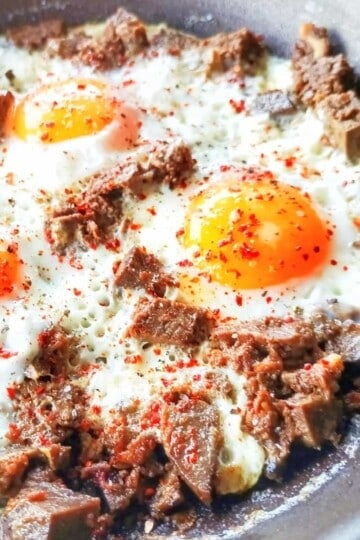 A close up of three fried eggs in a frying pan with chunks of beef scattered through them.