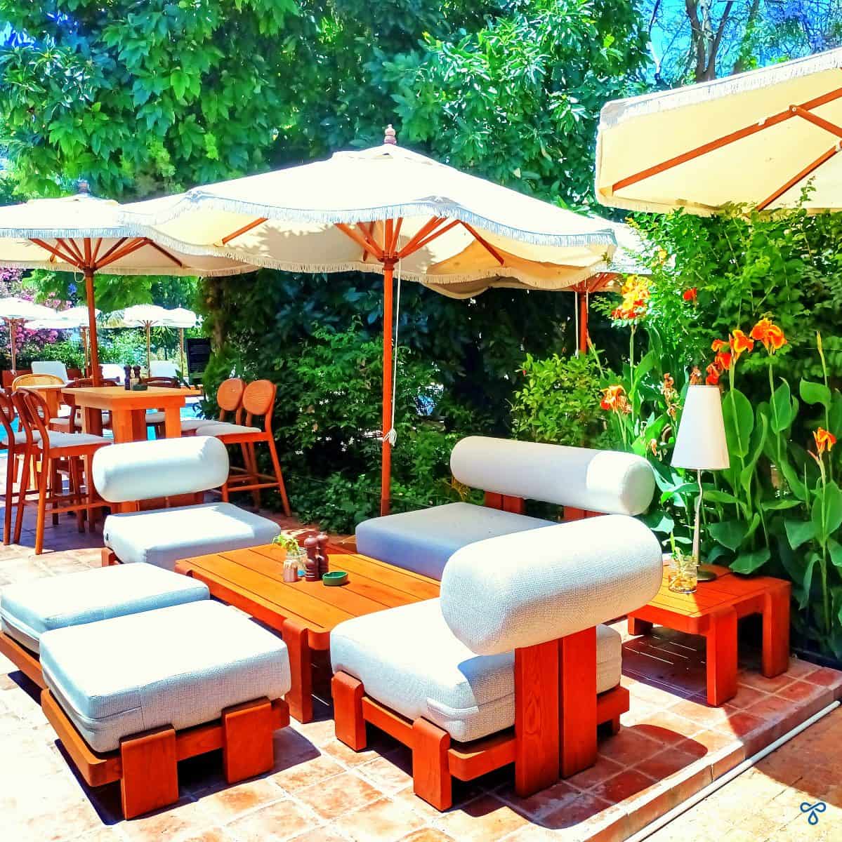 Wooden garden sofa and chairs with large cushions around a wooden coffee table, shaded by a parasol.