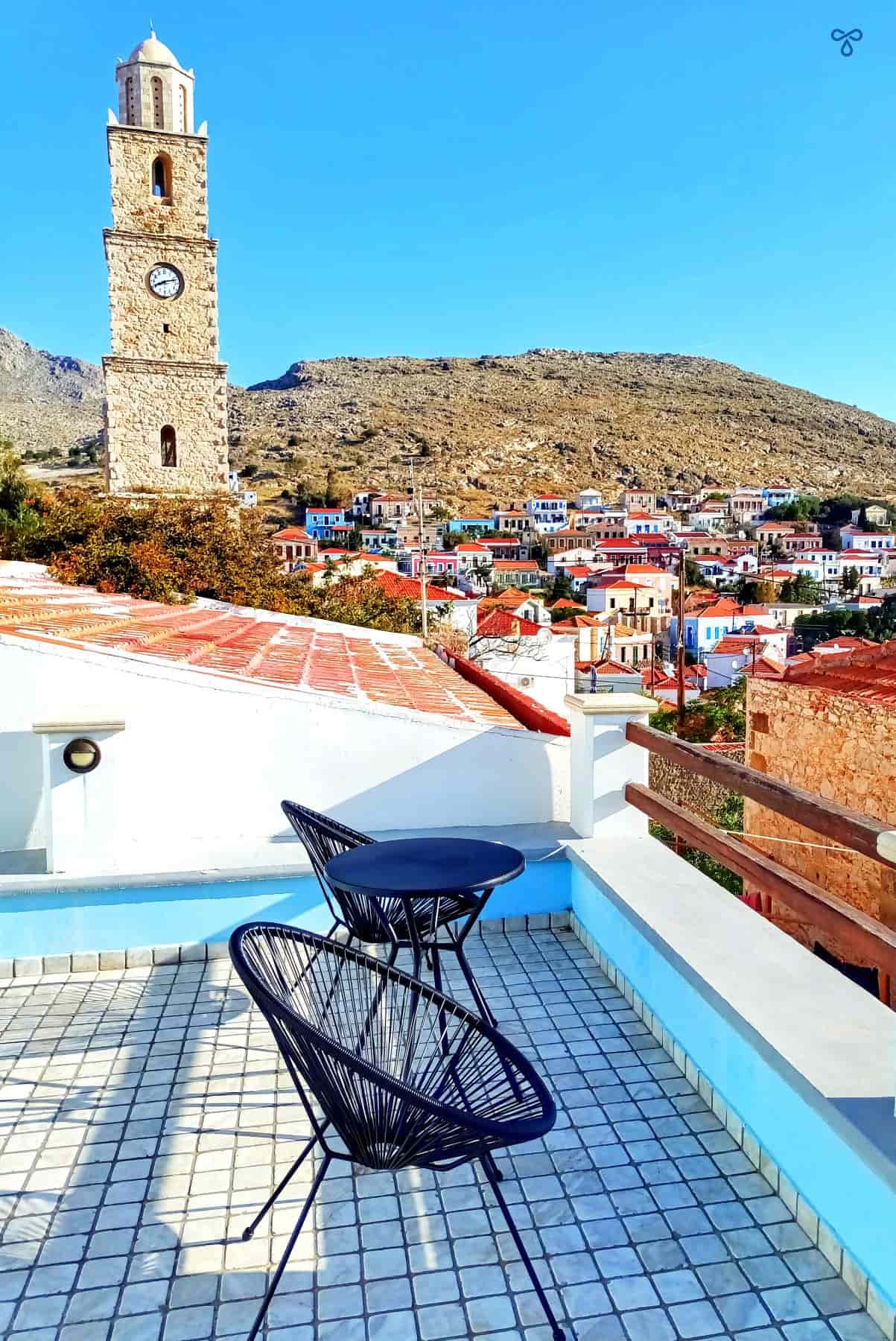Two chairs on a tiled terrace overlooking Halki houses on the hillside. The clocktower rises above the skyline.