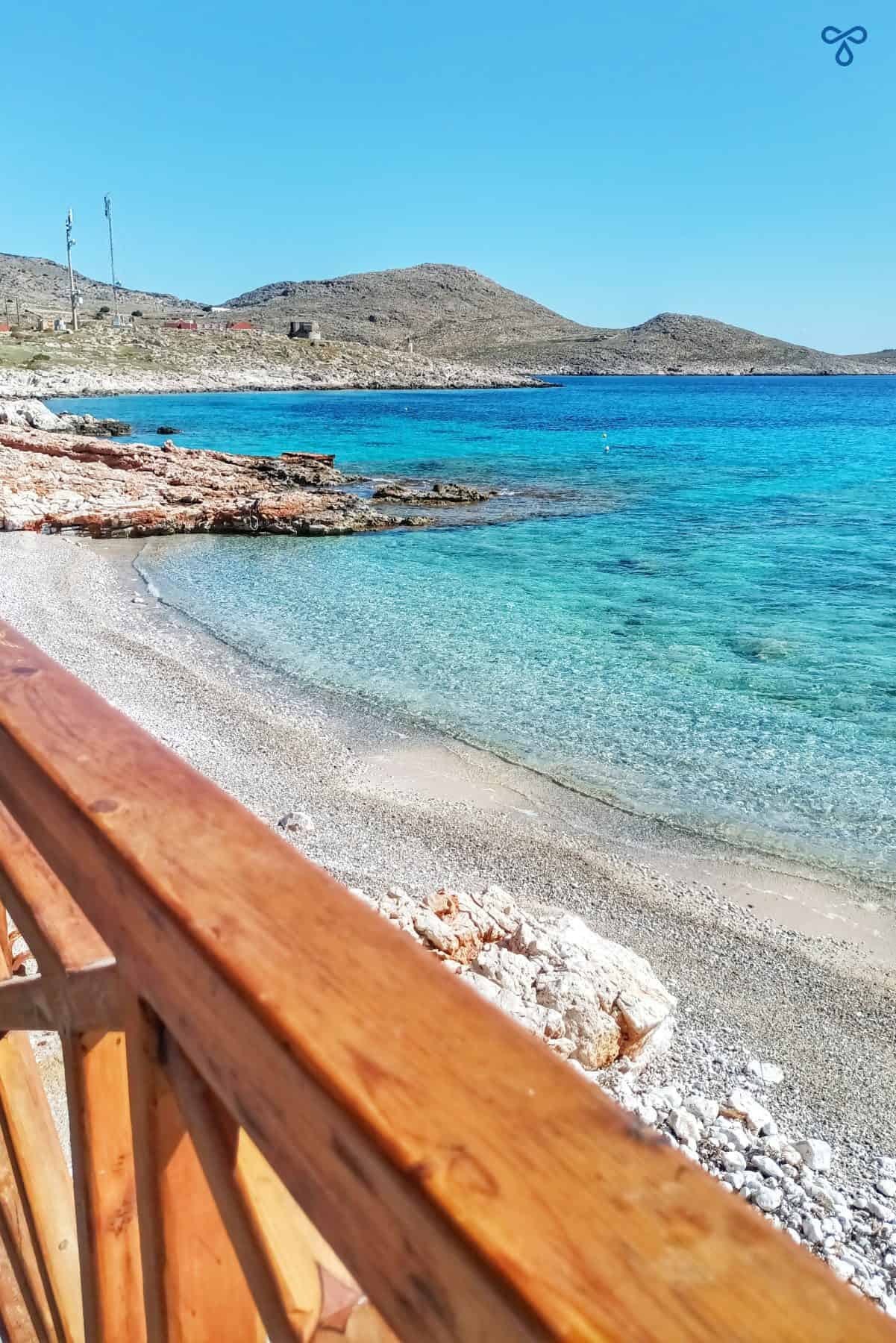 A wooden barrier leading to a stony beach and clear calm sea.