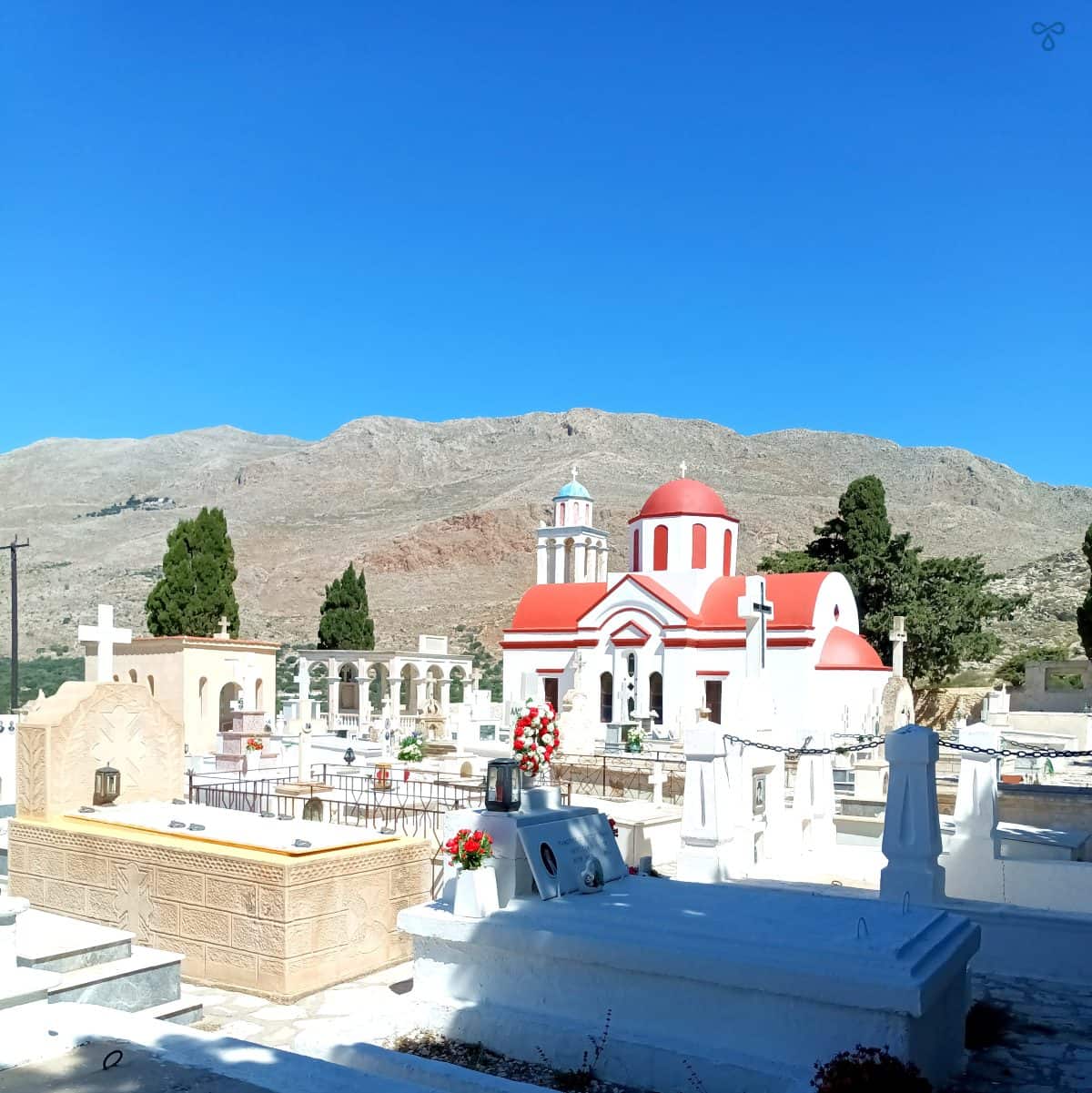 A white Greek chapel with a red domed roof and graves in the foreground.