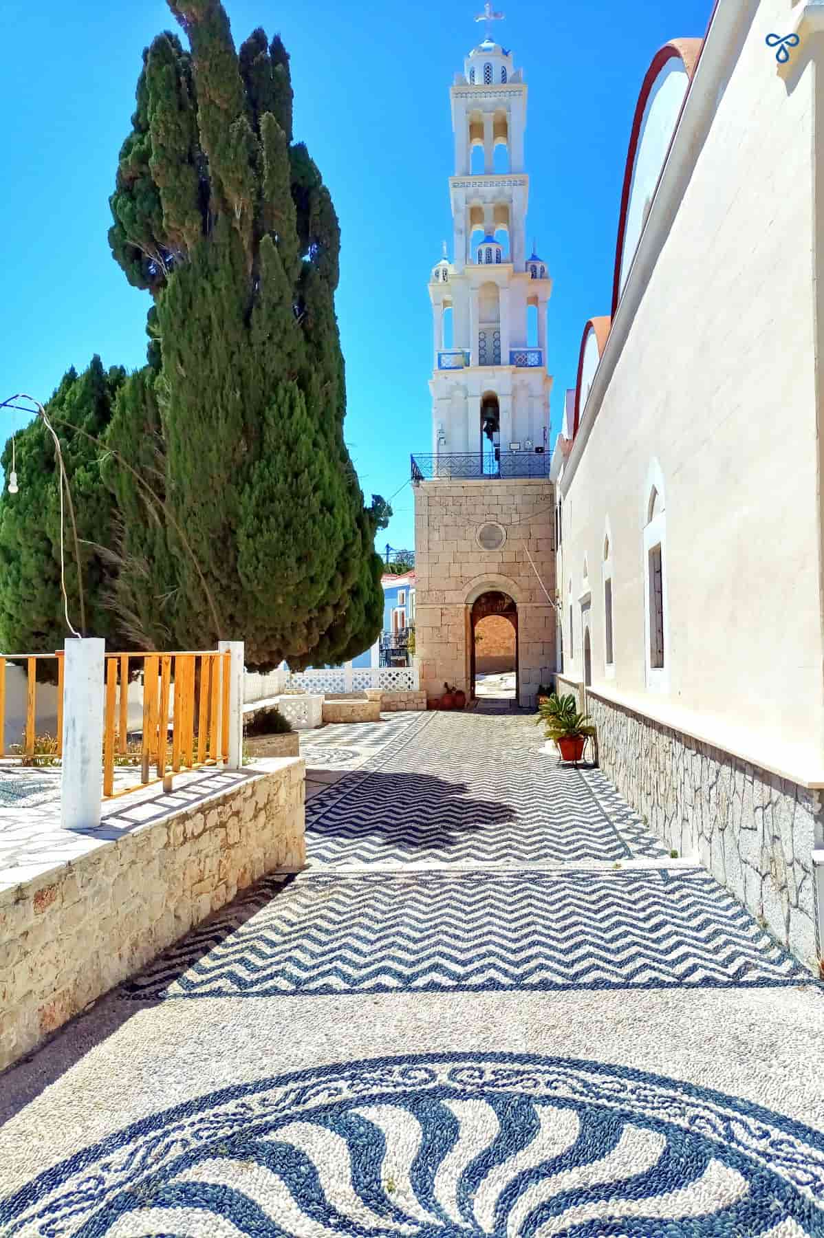 A tall white bell tower at the end of a church courtyard decorated with black and white pebble mosaics.