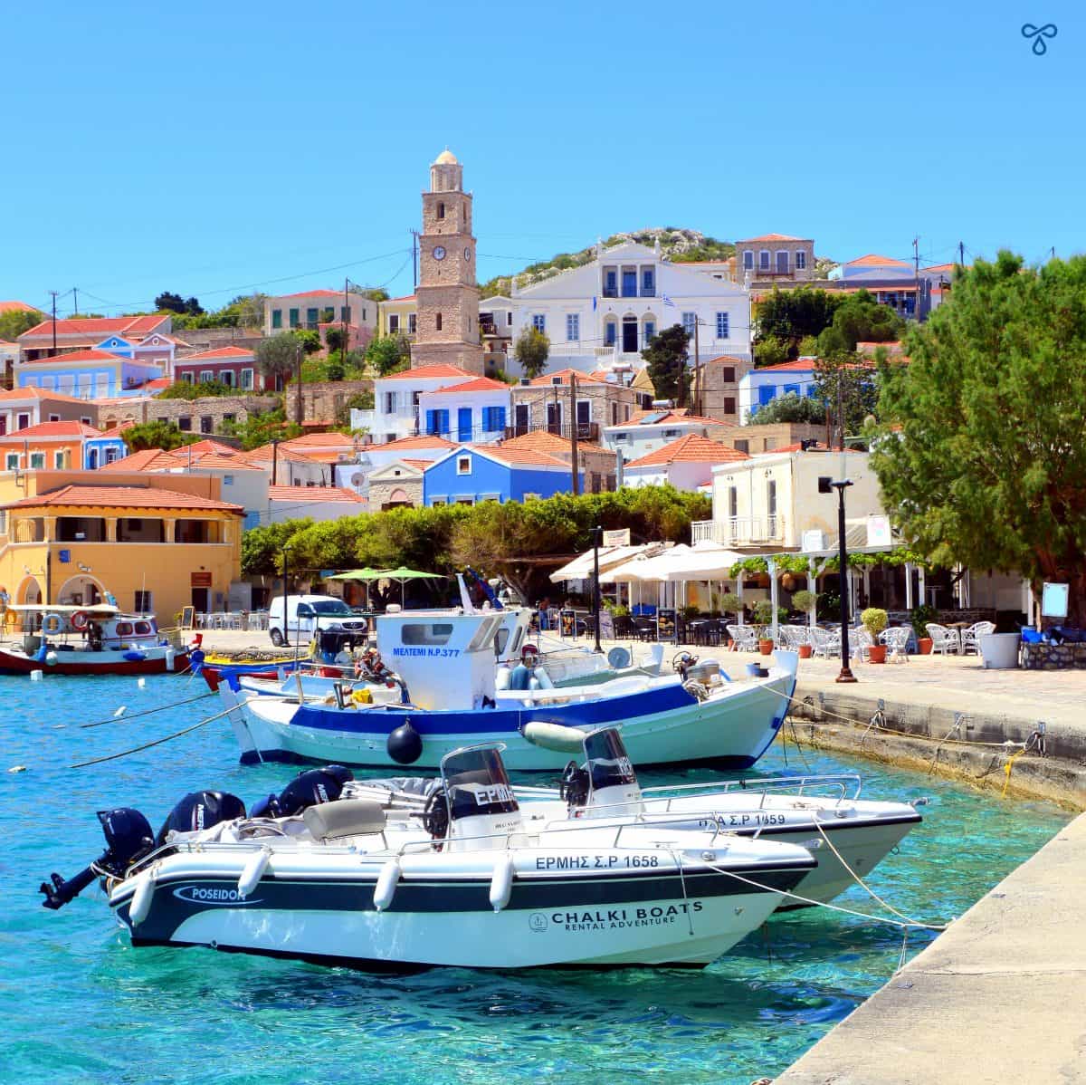 Small boats in Halki harbour with the clock tower rising above the colourful buildings behind.
