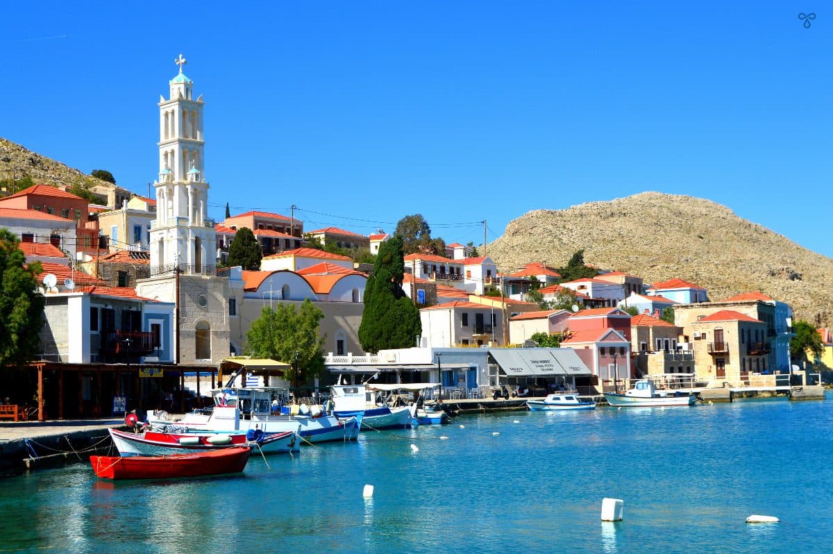 Looking over the harbour in Halki. The white bell tower of the church rises into the sky. Small fishing boats are tied up along the harbour wall.
