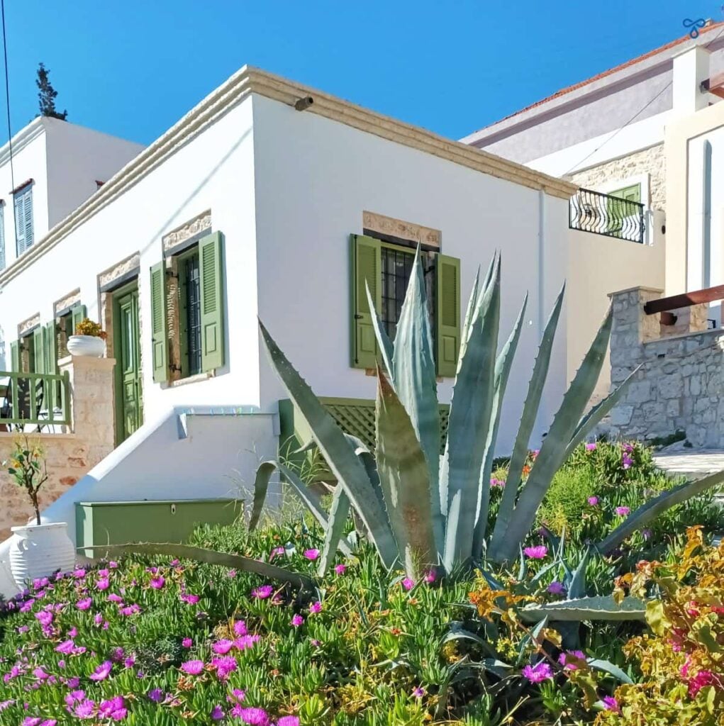 A whitewashed Greek house on Halki island with green window frames and shutters. A decorative garden is in the foreground.