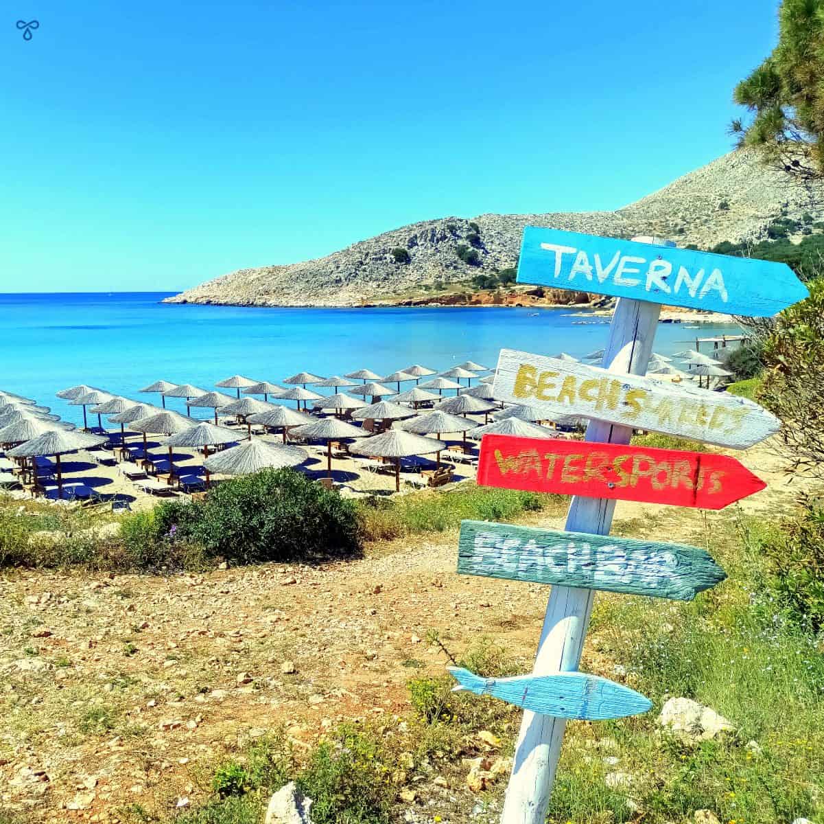 A wooden signpost with five arrows pointing to the local beach and taverna. The sea and beach umbrellas are in the background.