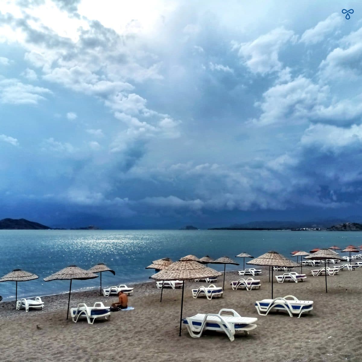 A grey, cloudy Çalış Beach with empty white sun loungers and rattan parasols on the beach.