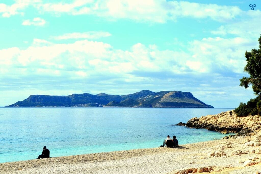Three people sitting on a pebble beach in Kaş overlooking the Greek island of Meis in the near distance.