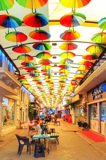 A pedestrianised shopping street covered by a canopy with multi-coloured umbrellas suspended from the canopy.