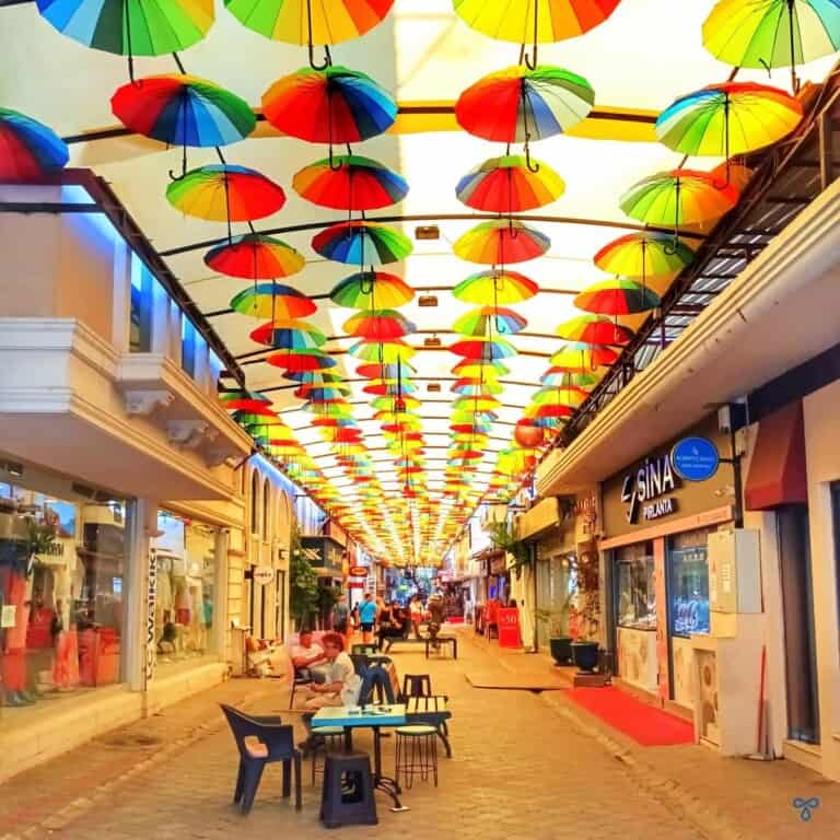 A pedestrianised shopping street covered by a canopy with multi-coloured umbrellas suspended from the canopy.