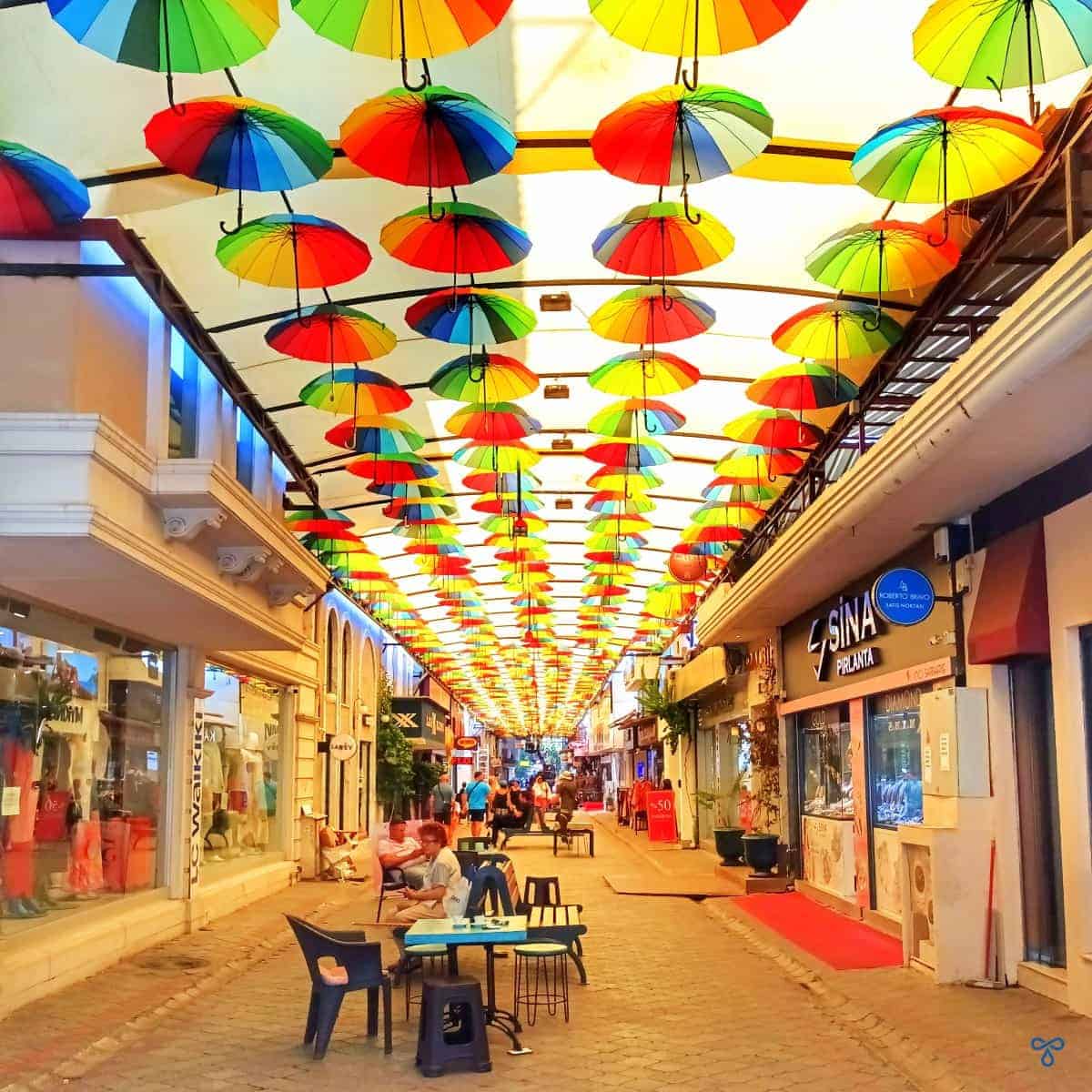A pedestrianised shopping street covered by a canopy with multi-coloured umbrellas suspended from the canopy.