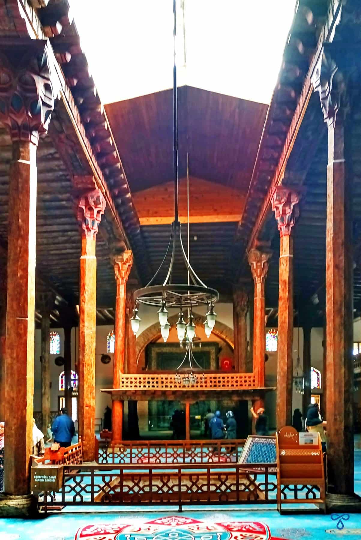 Looking down the centre of a mosque with wooden pillars either side. A large chandelier hangs from the ceiling and a skylight offers natural light.