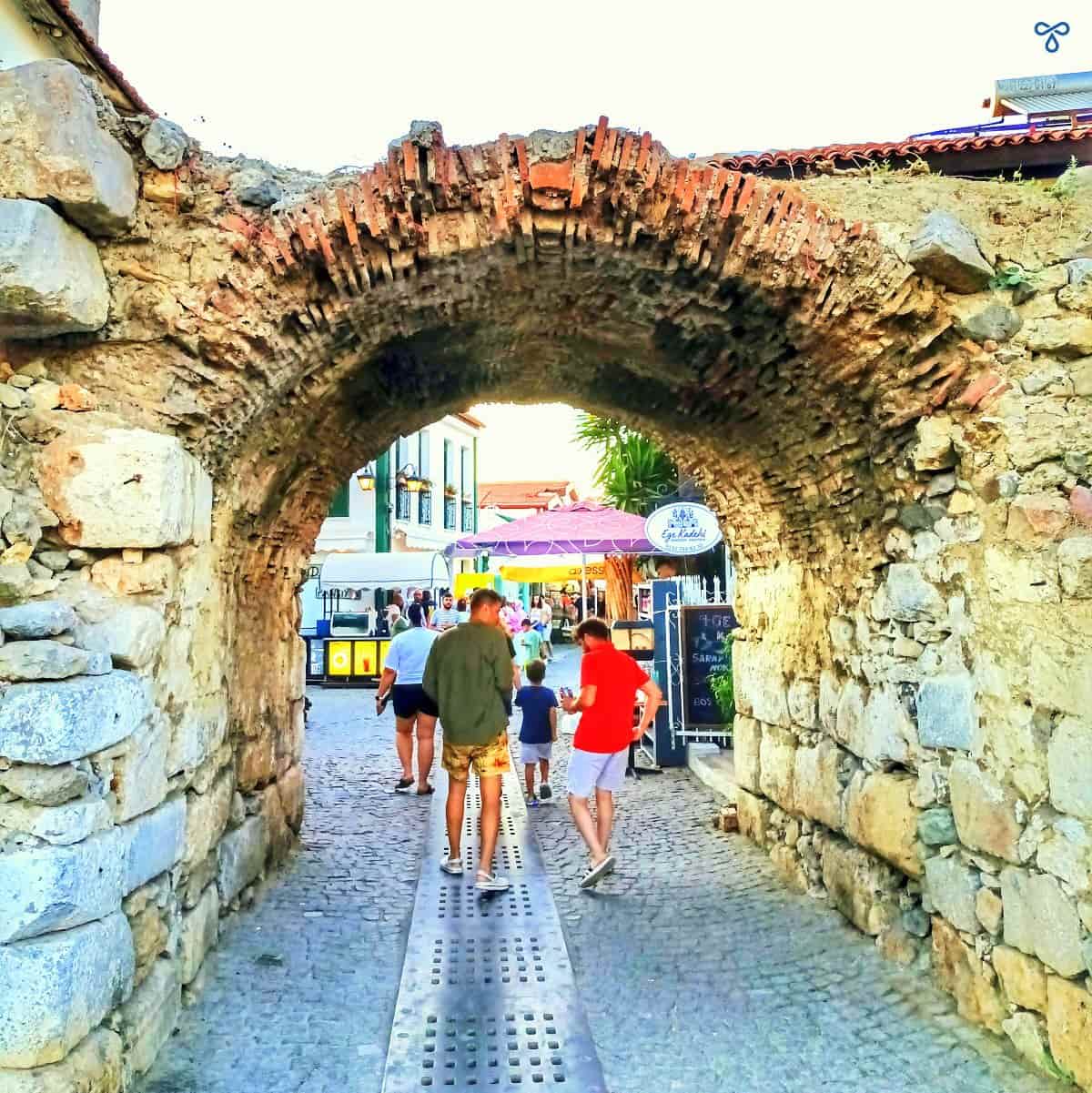 A historic arched stone entrance to Sığacık old town. Two people are walking through the arch.