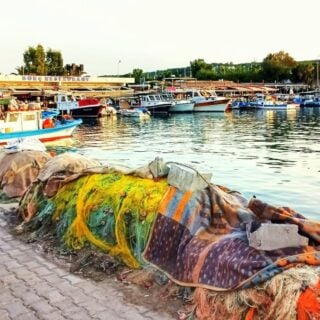 Colourful fishing nets line the curved water's edge in Sığacık harbour. Low rise restaurants and a small fishing boat are in the background.