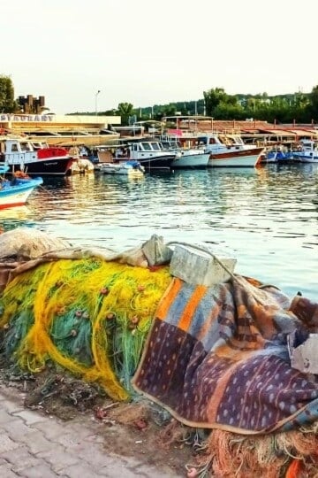 Colourful fishing nets line the curved water's edge in Sığacık harbour. Low rise restaurants and a small fishing boat are in the background.