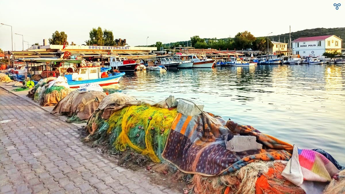 Colourful fishing nets line the curved water's edge in Sığacık harbour in Seferihisar. Low rise restaurants and a small fishing boat are in the background.