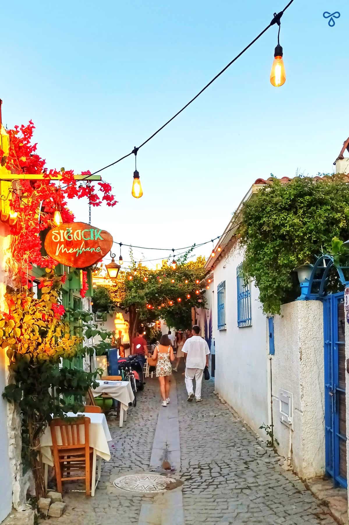 Golden hour, walking along narrow, cobbled alleyways in Sığacık. A string of bulbs hangs from walls either side of the pathway and bougainvillea climbs up the walls.