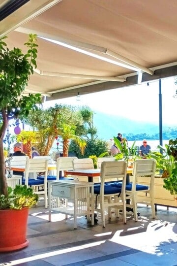 White tables and chairs shaded by an awning and framed by plants. There is a view of the sea outside.