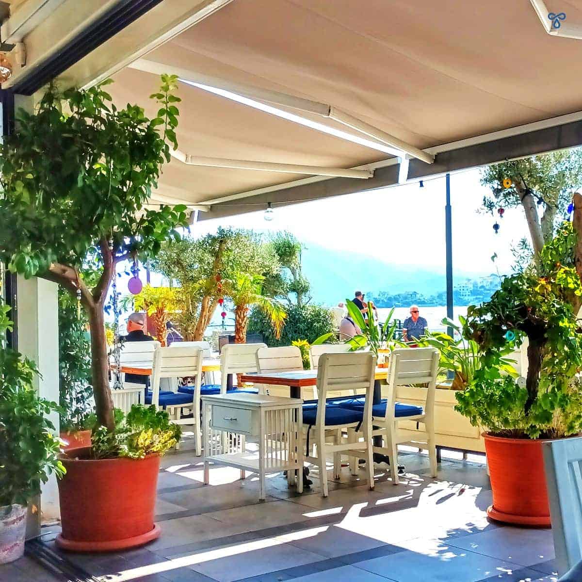 White tables and chairs shaded by an awning and framed by plants. There is a view of the sea outside Ju Restaurant.