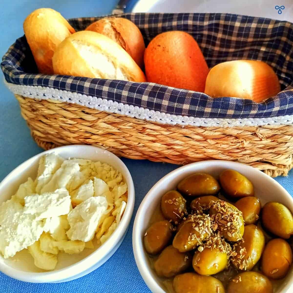 A basket of bread rolls and two small bowls filled with crumbled cheese and green olives.
