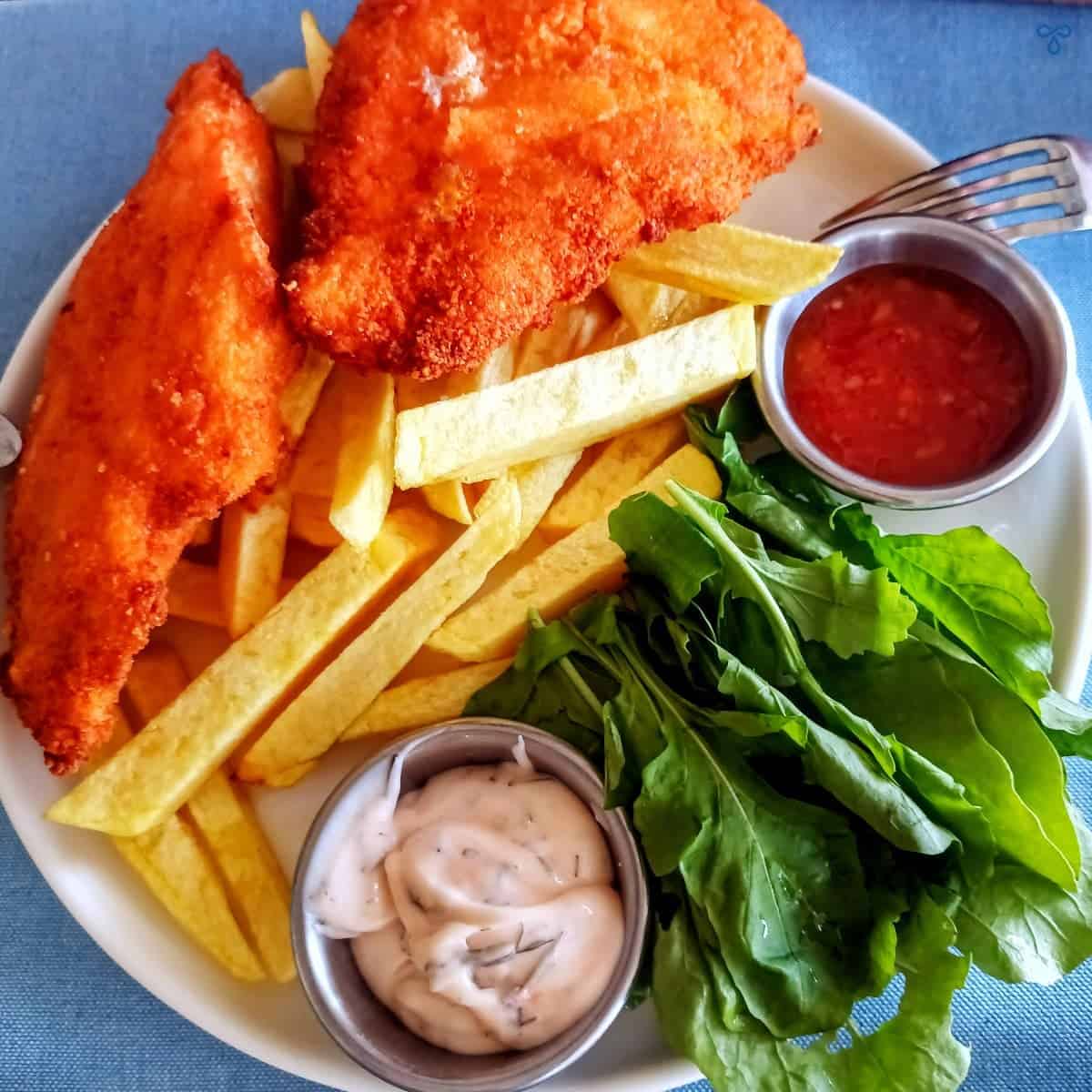 Two breaded chicken schnitzel on top of homemade chips and served with a side of rocket leaves and two dips.
